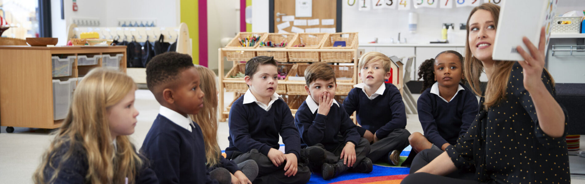 Female teacher holding up a book in front of her class of elementary school kids sitting on the floor in a classroom, side view