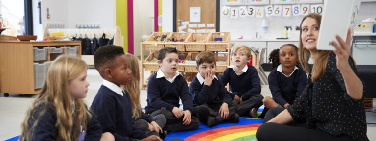 Female teacher holding up a book in front of her class of elementary school kids sitting on the floor in a classroom, side view