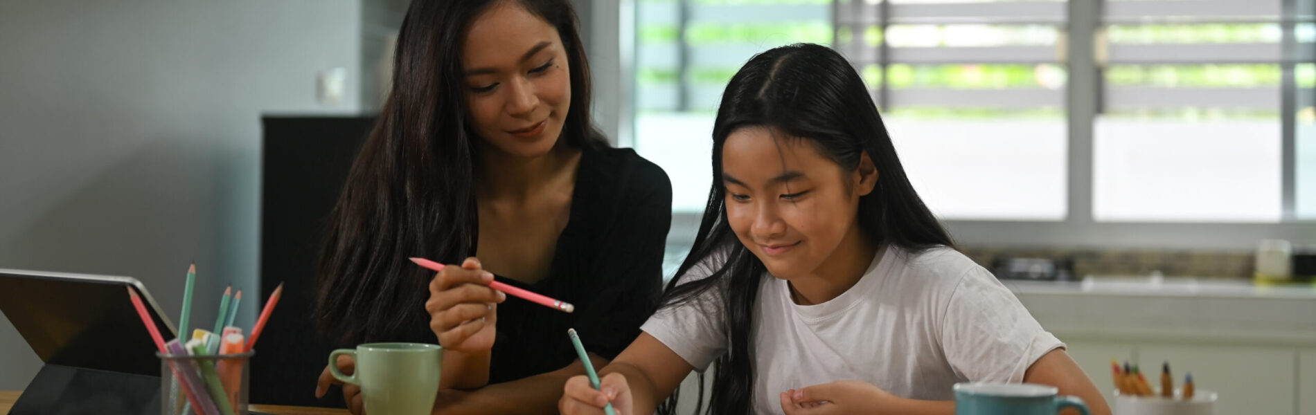 A mother is sitting with her daughter and teaching her homework at the wooden working desk.