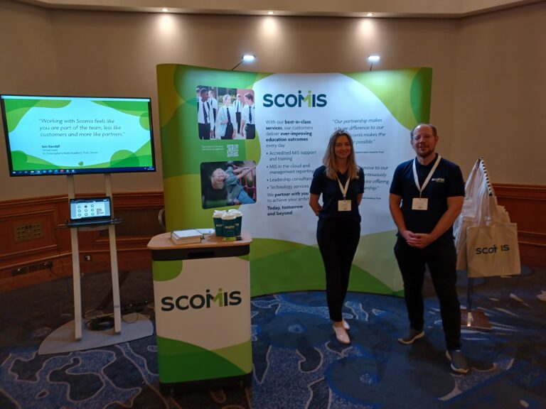 Photo of the ISBL Scomis stand with Amy and Michael standing in front of a large banner, screen displaying presentation and merch stand and bags, ready to greet existing and potential new customers